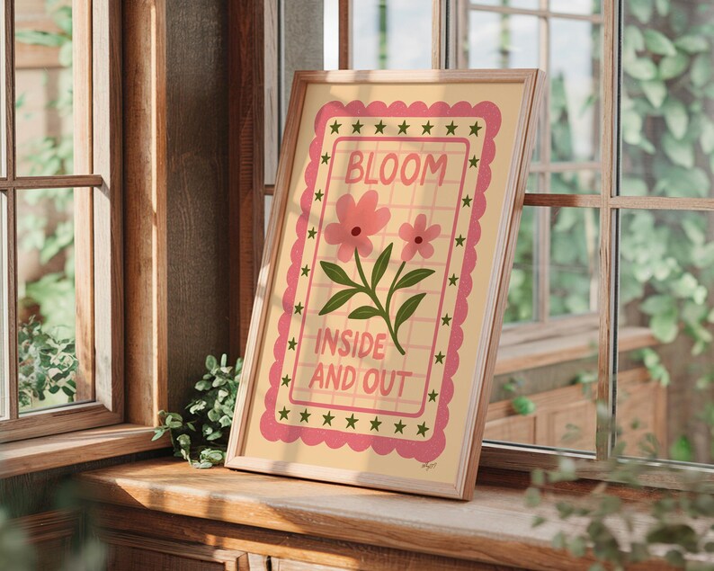 May include: A framed art print featuring the words "BLOOM INSIDE AND OUT" with pink flowers and green leaves. The artwork has a light yellow background with a pink border and star accents. The frame is a light wood color.