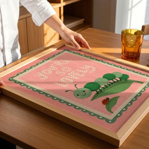 May include: A framed print with a pink background and the words "You are loved" in white. The print features a green caterpillar, a leaf, and a ladybug. The frame is made of light-colored wood.