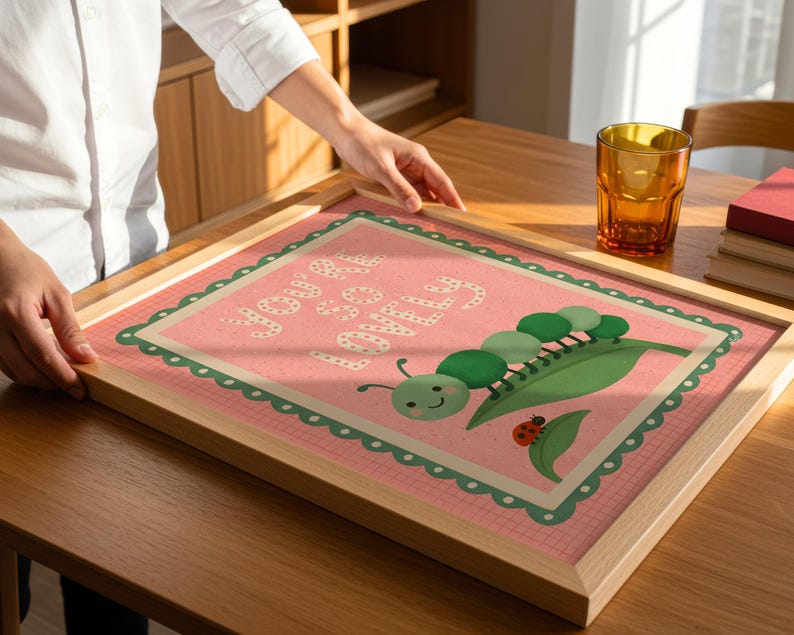 May include: A framed print with a pink background and the words "You are loved" in white. The print features a green caterpillar, a leaf, and a ladybug. The frame is made of light-colored wood.