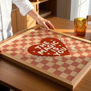 May include: A framed print featuring a red and cream chequered pattern and a red heart with the text "THE MAGIC IS YOU." The print is in a light wood frame, on a wooden table.