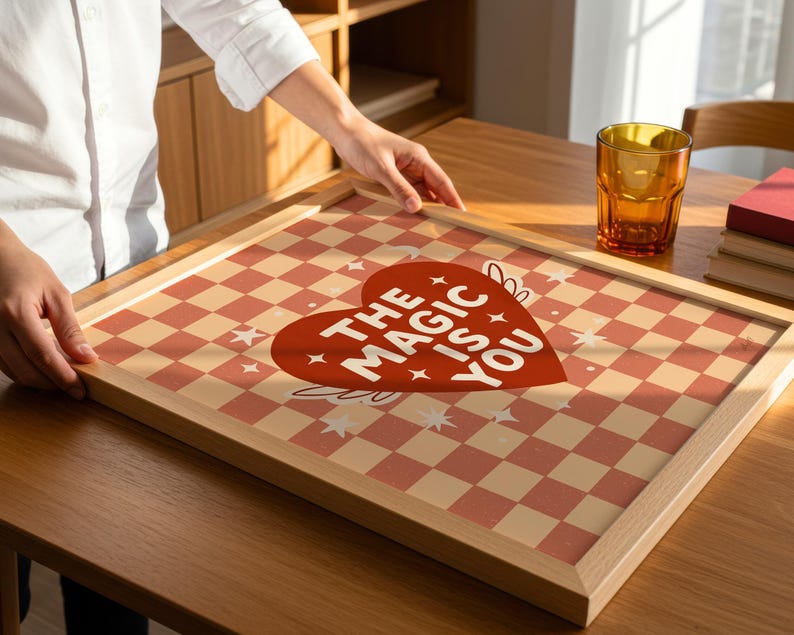 May include: A framed print featuring a red and cream chequered pattern and a red heart with the text "THE MAGIC IS YOU." The print is in a light wood frame, on a wooden table.
