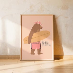 Puede incluir: Impresión enmarcada con un oso de dibujos animados con gorra rosa y pantalones cortos a cuadros, sosteniendo una tabla de surf. La impresión tiene el texto "Ride the wave of life with a big smile" sobre fondo rosa.