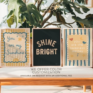 May include: Three rectangular wall art prints in light wood frames. The left print features yellow stripes, a blue scalloped border, and the text "You are my Sunshine". The center print is black with the text "SHINE BRIGHT". The right print has blue stripes and the text "you are the sweetest melody".