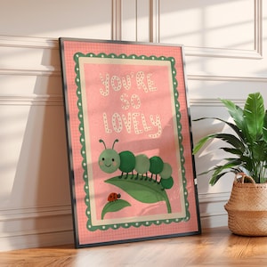 May include: A framed art print with the words "YOU'RE SO LOVELY" in white on a pink background. The print features a green caterpillar, a ladybug, and a green leaf. The frame is black.