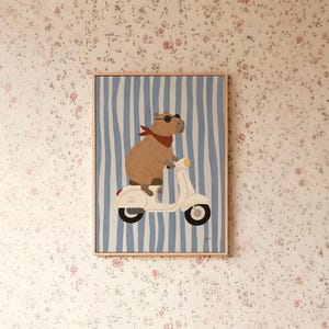 Pode incluir: Impress&atilde;o de arte emoldurada com uma capivara usando &oacute;culos de sol e um len&ccedil;o vermelho, andando de scooter branca em um fundo listrado azul e branco. A obra de arte est&aacute; em uma moldura de madeira e &eacute; exibida em papel de parede floral.