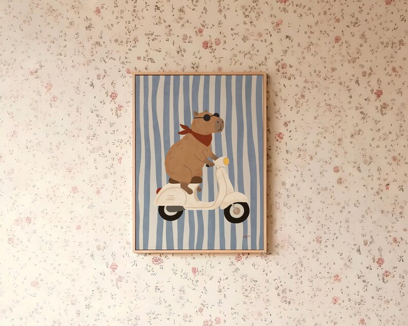 Pode incluir: Impress&atilde;o de arte emoldurada com uma capivara usando &oacute;culos de sol e um len&ccedil;o vermelho, andando de scooter branca em um fundo listrado azul e branco. A obra de arte est&aacute; em uma moldura de madeira e &eacute; exibida em papel de parede floral.