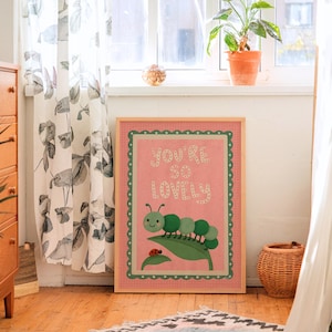 May include: A framed print with the text "YOU'RE SO LOVELY" in white letters on a pink background. The print features a green caterpillar on a leaf with a ladybug. The frame is light brown with a green scalloped border. The print is displayed in a room with a wooden floor.
