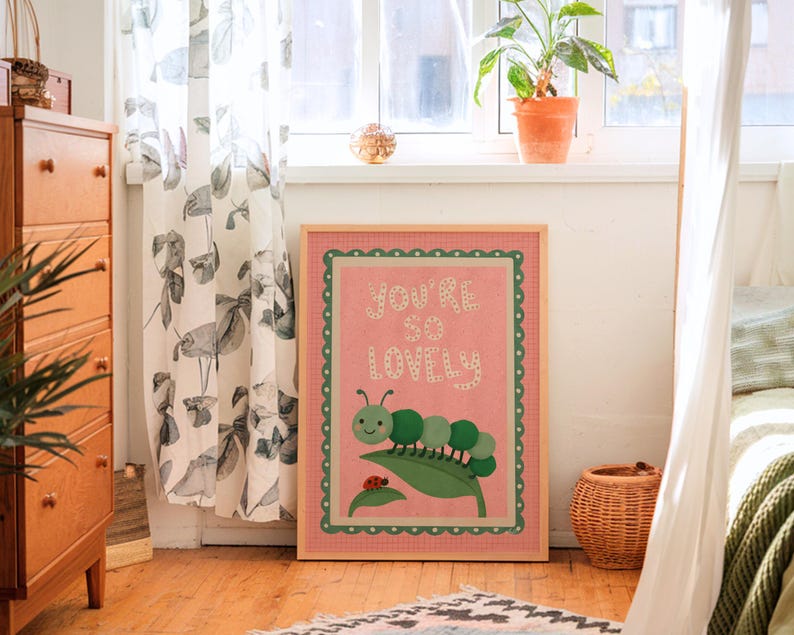 May include: A framed print with the text "YOU'RE SO LOVELY" in white letters on a pink background. The print features a green caterpillar on a leaf with a ladybug. The frame is light brown with a green scalloped border. The print is displayed in a room with a wooden floor.