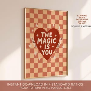 May include: A framed art print with a gold border. The print features a red heart with the words "THE MAGIC IS YOU" and small white wings. The background is a checkerboard pattern in shades of peach and red. The text "LOOKING FOR A DIFFERENT SIZE OR COLOR? SEND US A MESSAGE!" is also included.
