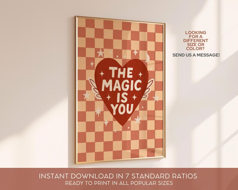 May include: A framed art print with a gold border. The print features a red heart with the words "THE MAGIC IS YOU" and small white wings. The background is a checkerboard pattern in shades of peach and red. The text "LOOKING FOR A DIFFERENT SIZE OR COLOR? SEND US A MESSAGE!" is also included.