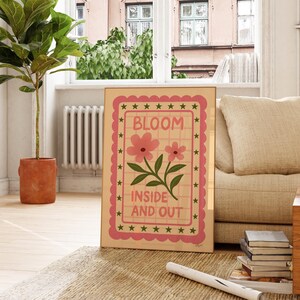 May include: A framed art print with the words "BLOOM INSIDE AND OUT" in a pink and green design. The artwork features pink flowers and green leaves on a beige background, framed with a pink border and green stars. The print is displayed indoors.