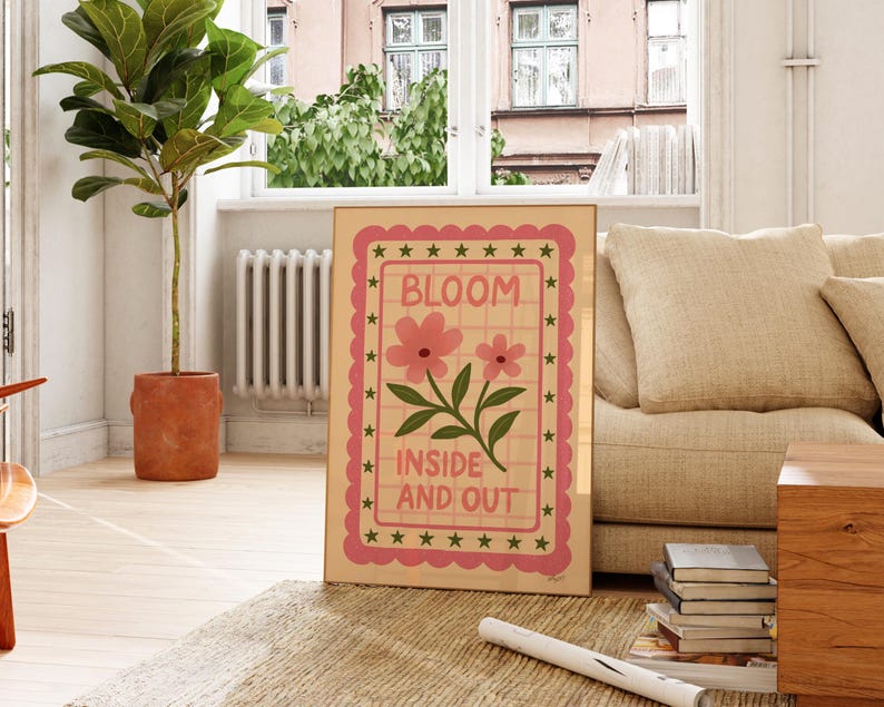 May include: A framed art print with the words "BLOOM INSIDE AND OUT" in a pink and green design. The artwork features pink flowers and green leaves on a beige background, framed with a pink border and green stars. The print is displayed indoors.