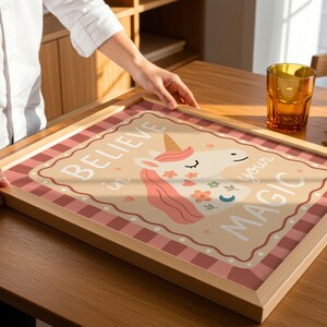 May include: A framed print with a unicorn illustration and the words "Believe in your Magic." The artwork features a light brown background with a pink and white checkered border. The unicorn has a pink mane and floral accents.