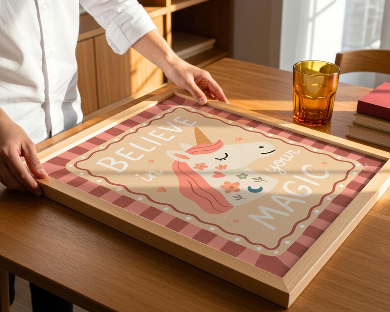May include: A framed print with a unicorn illustration and the words "Believe in your Magic." The artwork features a light brown background with a pink and white checkered border. The unicorn has a pink mane and floral accents.