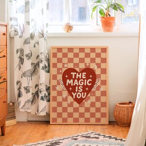 May include: A framed art print featuring a red and tan checkered pattern. The print has a red heart with white text that reads "THE MAGIC IS YOU". The heart has white wings and is surrounded by stars.