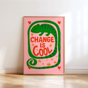 Puede incluir: Una impresión artística enmarcada con un fondo rosa presenta un camaleón verde y las palabras "CHANGE IS COOL" en rojo y verde. La impresión incluye pequeños corazones, estrellas y detalles florales. La obra de arte se exhibe sobre un suelo de madera.