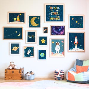 Puede incluir: Una pared blanca con una galería de impresiones enmarcadas con ilustraciones de temática espacial. Las impresiones incluyen un cohete, planetas, estrellas, astronautas y una galaxia. El texto en una de las impresiones dice "Reach for the Stars, Never Stop Dreaming!"