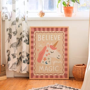 May include: A framed print with a unicorn illustration and the words "BELIEVE in your MAGIC". The unicorn has a pink mane and a golden horn, with floral and crescent moon accents. The frame has a pink and white checkered border.