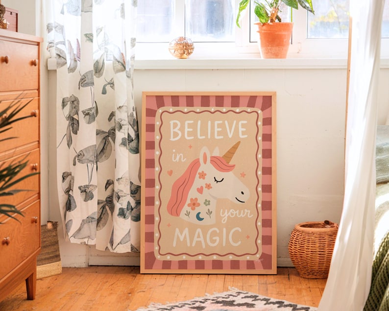 May include: A framed print with a unicorn illustration and the words "BELIEVE in your MAGIC". The unicorn has a pink mane and a golden horn, with floral and crescent moon accents. The frame has a pink and white checkered border.