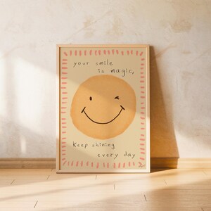 Puede incluir: Una impresión enmarcada con un fondo beige claro presenta una gran carita sonriente guiñando un ojo. El texto "your smile is magic, keep shining every day" está escrito arriba y abajo de la carita sonriente. El marco es marrón claro.
