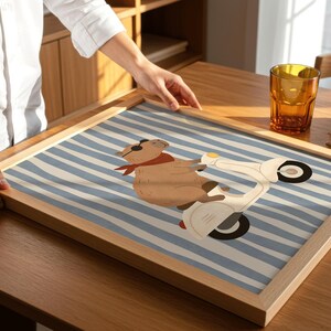 Pode incluir: Uma impress&atilde;o art&iacute;stica emoldurada com um capivara de desenho animado em uma scooter branca contra um fundo listrado azul e branco. O capivara usa um len&ccedil;o vermelho e &oacute;culos de sol. A moldura &eacute; de madeira clara.