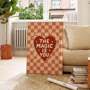 May include: A framed art print featuring a peach and cream checkerboard design. A red heart with wings and the words "THE MAGIC IS YOU" are displayed on the print. The print is placed on a rug near a sofa and a potted plant.