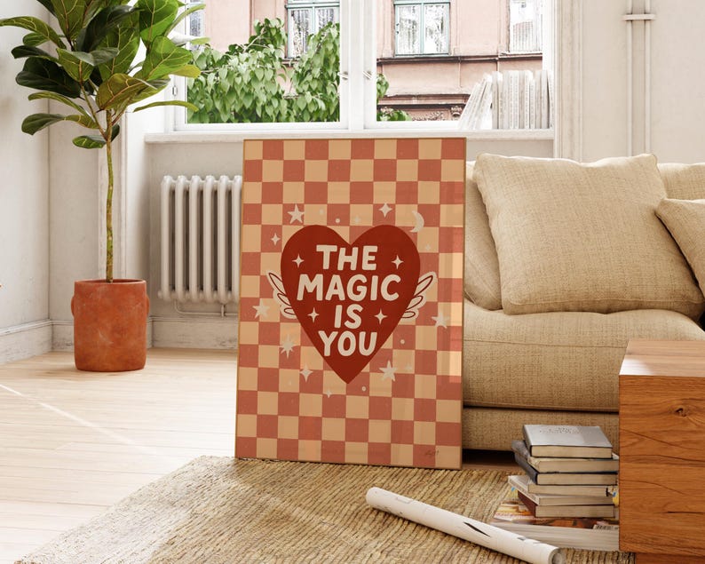 May include: A framed art print featuring a peach and cream checkerboard design. A red heart with wings and the words "THE MAGIC IS YOU" are displayed on the print. The print is placed on a rug near a sofa and a potted plant.