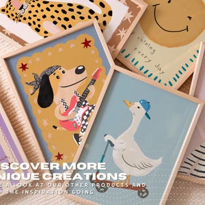 May include: A collection of framed art prints with whimsical animal illustrations. The prints feature a dog playing guitar, a goose on a scooter, and a smiling sun. The text "DISCOVER MORE UNIQUE CREATIONS" is visible.