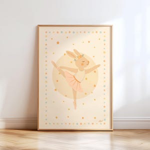 Puede incluir: Una impresión artística enmarcada que presenta un conejito bailando ballet con un tutú rosa y una corona de flores. La ilustración está ambientada sobre un fondo amarillo suave con estrellas y un borde punteado, creando una estética caprichosa y encantadora.