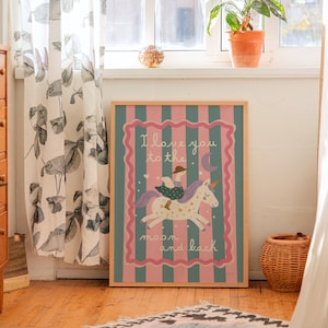 May include: A framed print with a pink and teal striped background features a whimsical illustration of a unicorn with a rider. The text "I love you to the moon and back" is displayed in a decorative font. The artwork is set in a room with natural light.