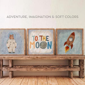 May include: Three framed illustrations with a space theme. The first shows a brown bear wearing an astronaut suit. The second shows a moon with the text "To the Moon". The third shows a rocket with a white cat inside. The illustrations are on a wooden shelf.