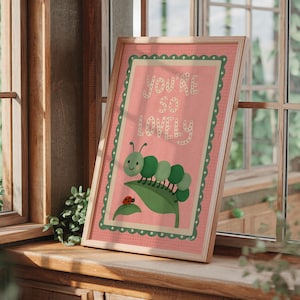 May include: Framed art print with a pink background and green border. The print features a green caterpillar, a red ladybug, and the words "YOU'RE SO LOVELY" in white lettering. The artwork is displayed on a wooden windowsill.