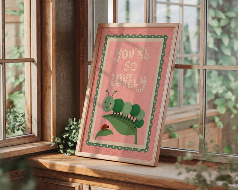 May include: Framed art print with a pink background and green border. The print features a green caterpillar, a red ladybug, and the words "YOU'RE SO LOVELY" in white lettering. The artwork is displayed on a wooden windowsill.