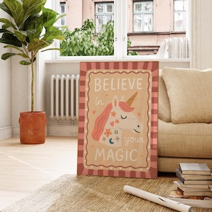 May include: A framed canvas print featuring a whimsical unicorn illustration with the text "BELIEVE in your MAGIC." The artwork has a tan background with a pink and red plaid border. The unicorn has a golden horn and pink mane.