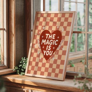 May include: A framed print with a red heart design and the words "THE MAGIC IS YOU". The background has a checkered pattern in orange and cream. The wooden framed print is displayed against a window, creating a warm, inviting aesthetic.