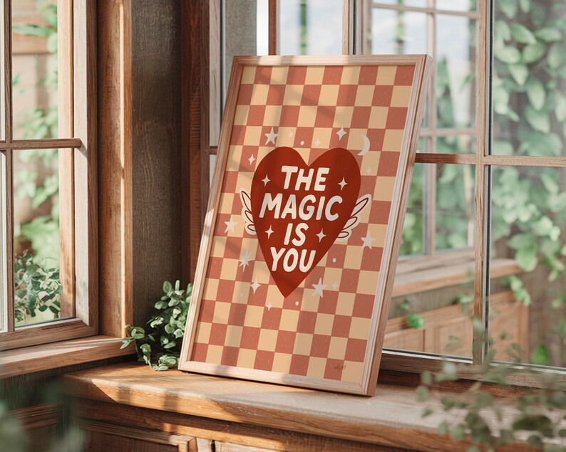 May include: A framed print with a red heart design and the words "THE MAGIC IS YOU". The background has a checkered pattern in orange and cream. The wooden framed print is displayed against a window, creating a warm, inviting aesthetic.