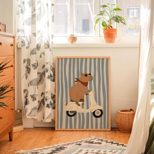 Pode incluir: Uma impress&atilde;o de arte emoldurada com uma capivara em uma scooter branca. A capivara usa &oacute;culos de sol e uma bandana vermelha em um fundo listrado azul e branco. A obra de arte &eacute; exibida em uma sala com uma c&ocirc;moda de madeira e uma janela.