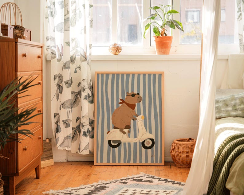 Pode incluir: Uma impress&atilde;o de arte emoldurada com uma capivara em uma scooter branca. A capivara usa &oacute;culos de sol e uma bandana vermelha em um fundo listrado azul e branco. A obra de arte &eacute; exibida em uma sala com uma c&ocirc;moda de madeira e uma janela.
