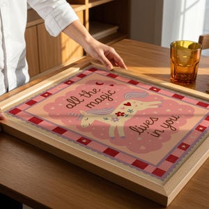 May include: A framed print with a light wood frame. The print features a unicorn illustration with the text "all the magic lives in you" in a pink and red color scheme. The print is on a wooden table.