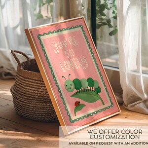 May include: A framed print with a pink background and the words "You're so Lovely" in white. The print features a green caterpillar on a leaf with a ladybug. The frame is gold, and the print is leaning against a woven basket.