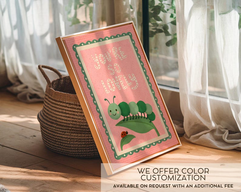 May include: A framed print with a pink background and the words "You're so Lovely" in white. The print features a green caterpillar on a leaf with a ladybug. The frame is gold, and the print is leaning against a woven basket.