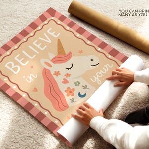 May include: A rolled-up poster featuring a unicorn illustration with the words "BELIEVE in your" in white. The unicorn has a pink mane and floral accents. The poster is on a light-colored surface.