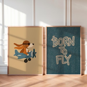 May include: Two framed wall art prints. The left print features a brown dog wearing a brown aviator cap and goggles, piloting a blue airplane against a tan background. The right print displays the text BORN TO FLY in tan on a dark teal background.