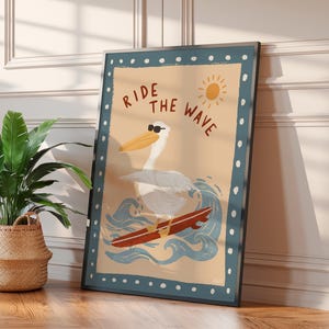 May include: A framed art print featuring a pelican wearing sunglasses surfing on a red surfboard. The artwork has a light blue border with white dots and the words "RIDE THE WAVE" in brown. A sun illustration is in the upper right corner.