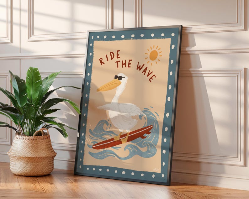 May include: A framed art print featuring a pelican wearing sunglasses surfing on a red surfboard. The artwork has a light blue border with white dots and the words "RIDE THE WAVE" in brown. A sun illustration is in the upper right corner.