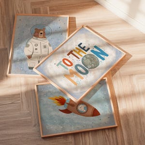 May include: Three framed art prints with a space theme. One print features a bear in a spacesuit, another says "TO THE MOON", and the third shows a rocket ship with a bear inside. The prints have a light blue background with a wooden frame.