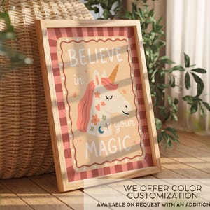 May include: A framed print featuring a whimsical unicorn illustration. The artwork has a light brown background with the words "BELIEVE in your MAGIC" and a unicorn with a pink mane and golden horn. The frame is light wood. The print is on a wooden surface.