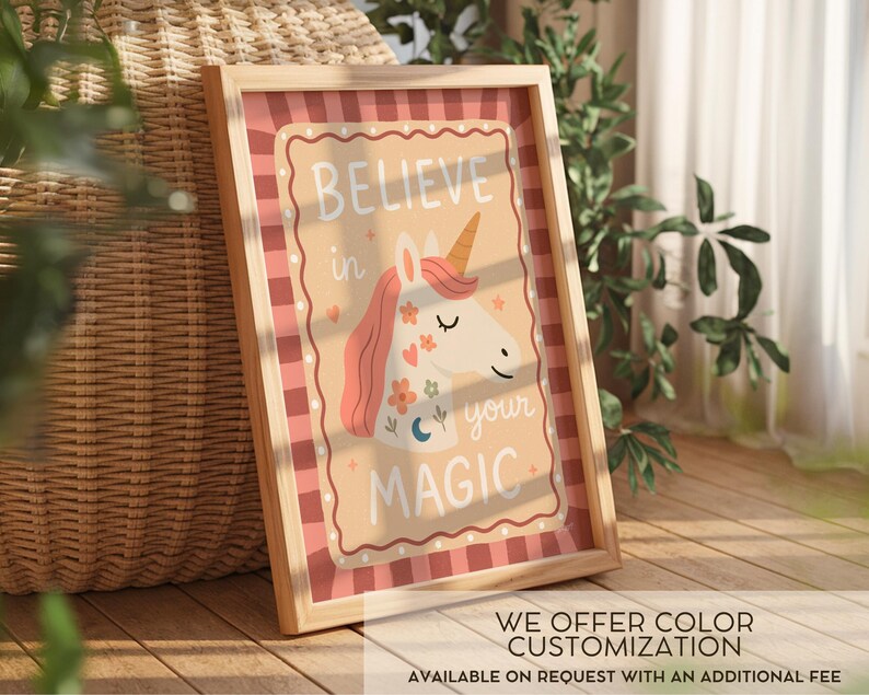 May include: A framed print featuring a whimsical unicorn illustration. The artwork has a light brown background with the words "BELIEVE in your MAGIC" and a unicorn with a pink mane and golden horn. The frame is light wood. The print is on a wooden surface.