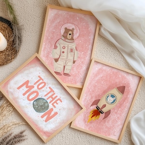 May include: Three framed art prints with a space theme. One print features a bear in a spacesuit, another says "TO THE MOON" with a planet, and the third shows a rocket ship with a bunny. The prints have a pink background and wooden frames.