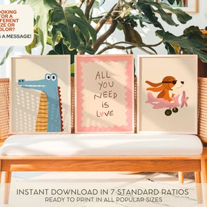 Op de afbeelding: Drie ingelijste kunstafdrukken met cartoonillustraties. E&eacute;n toont een blauwe alligator, een andere heeft de tekst "ALL YOU NEED IS LOVE", en de derde toont een hond in een vliegtuig. De prints worden op een houten bank getoond.